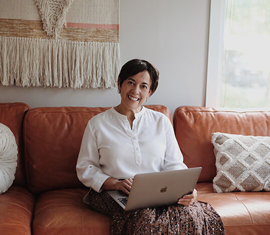 A smiling 65 year old woman with short hair sitting on the couch of a cozy living room with a laptop on her lap.